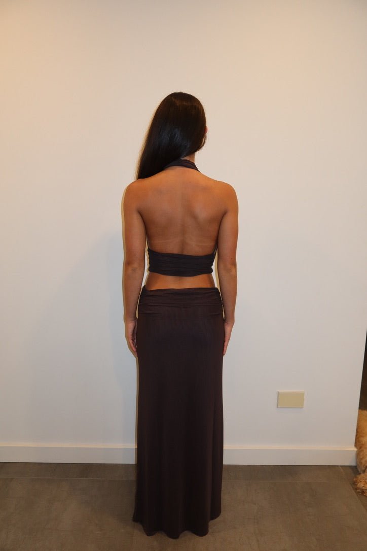 A woman viewed from behind wearing a dark brown halter-neck maxi dress with a wrap-style back cutout, standing against a plain off-white wall.