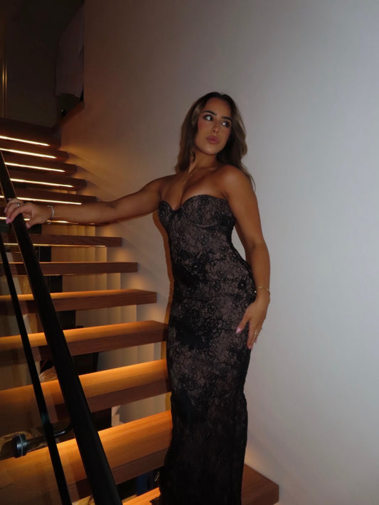 A woman wearing a strapless black lace evening gown stands on a wooden staircase indoors, holding the handrail.