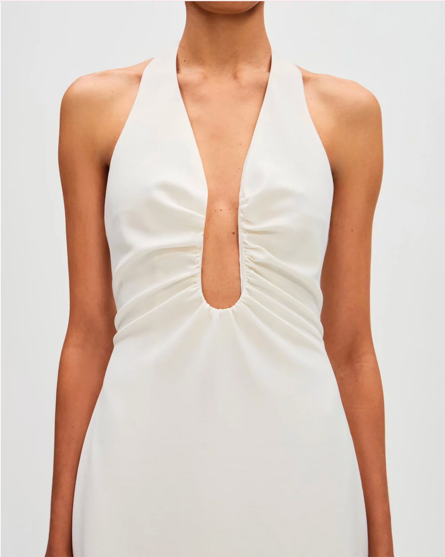 A close-up of a model wearing a white halter-neck dress with a deep plunging V-neckline and gathered ruching around the center front cutout.
