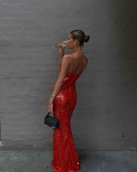A woman in a shimmering red sequined mermaid gown drinks from a glass while holding a small black clutch against a gray textured wall.