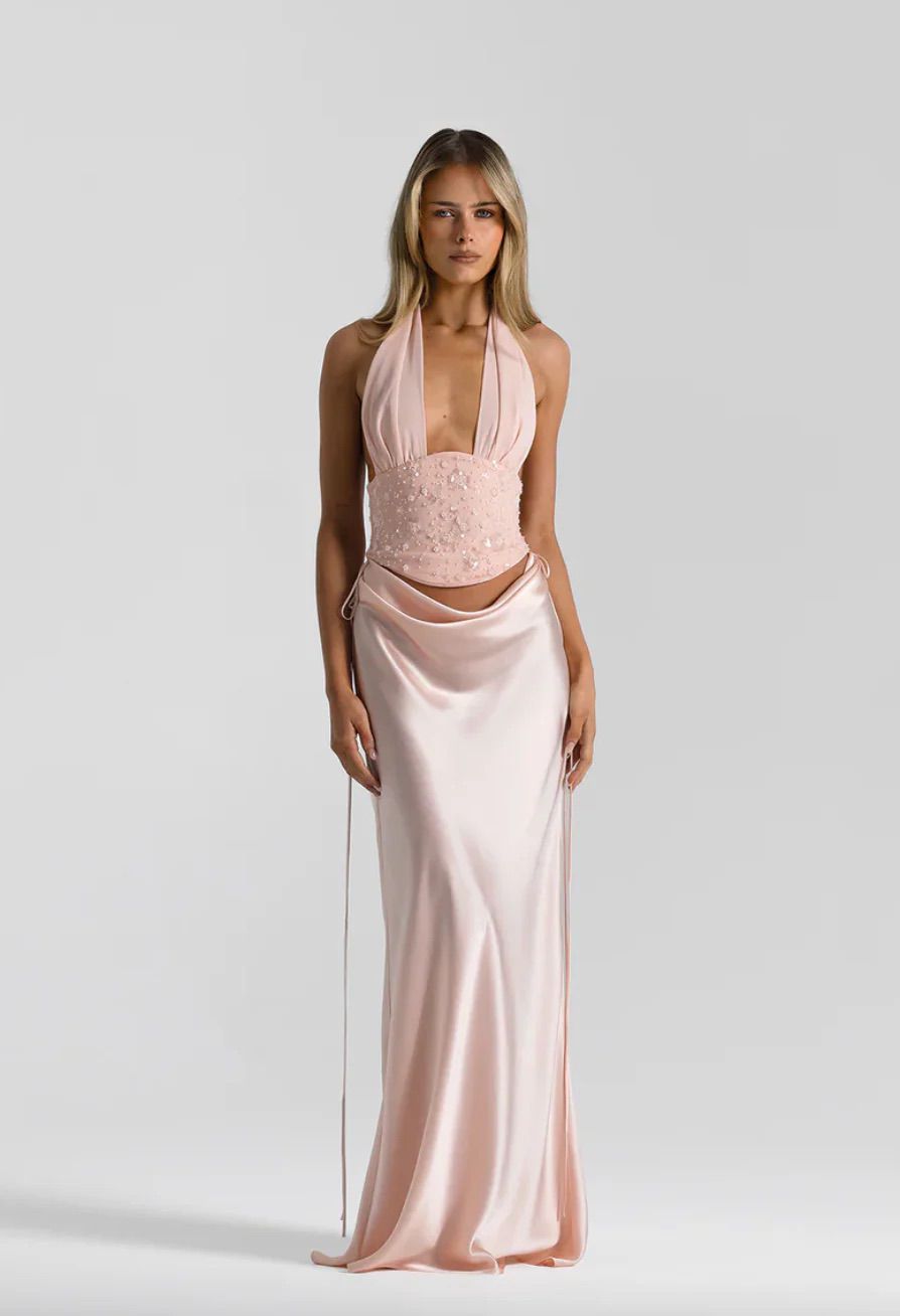 A model wearing a pale pink halter-neck evening gown with a lace-embellished bodice and a satin-like flowing skirt, photographed against a light gray studio background.
