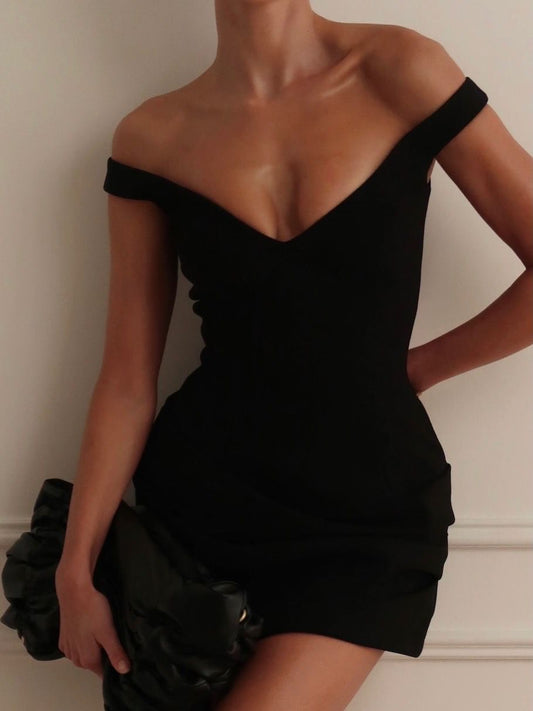 A woman wearing a fitted off-the-shoulder black mini dress, holding a black ruffled clutch against a light beige wall.