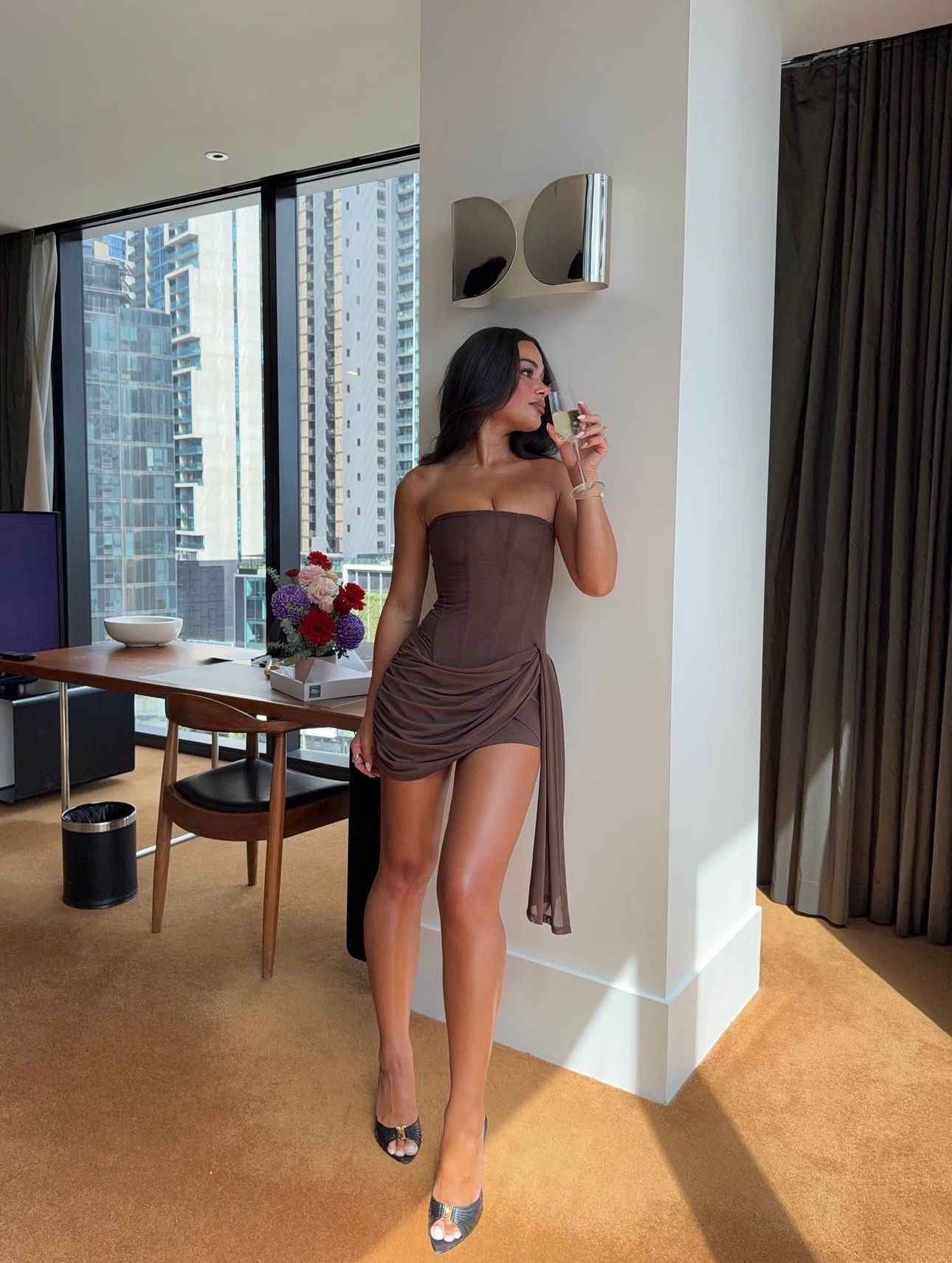 A woman wearing a brown strapless ruched mini dress and silver heels stands in a modern room with a large window showing city buildings outside.