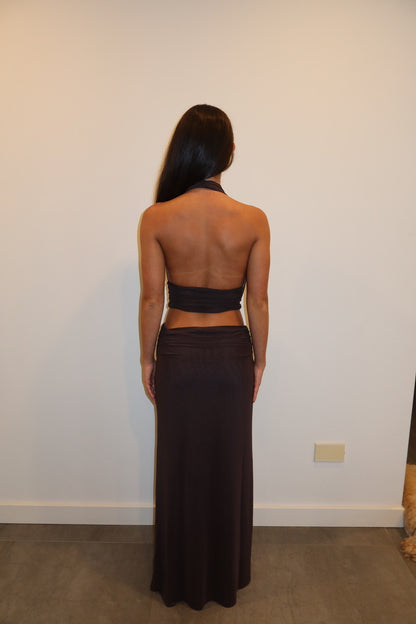 A woman viewed from behind wearing a dark brown halter-neck maxi dress with a wrap-style back cutout, standing against a plain off-white wall.