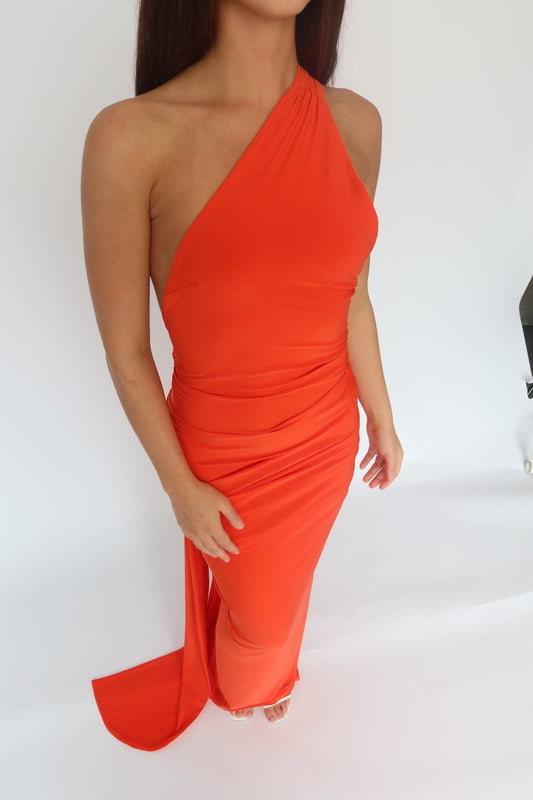 A woman modeling a bright orange-red one-shoulder draped dress against a plain white studio background.