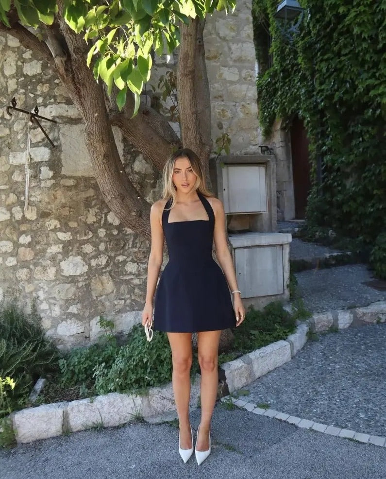 A woman wearing a navy halter mini dress and white pointed heels stands outdoors by a stone wall and tree.