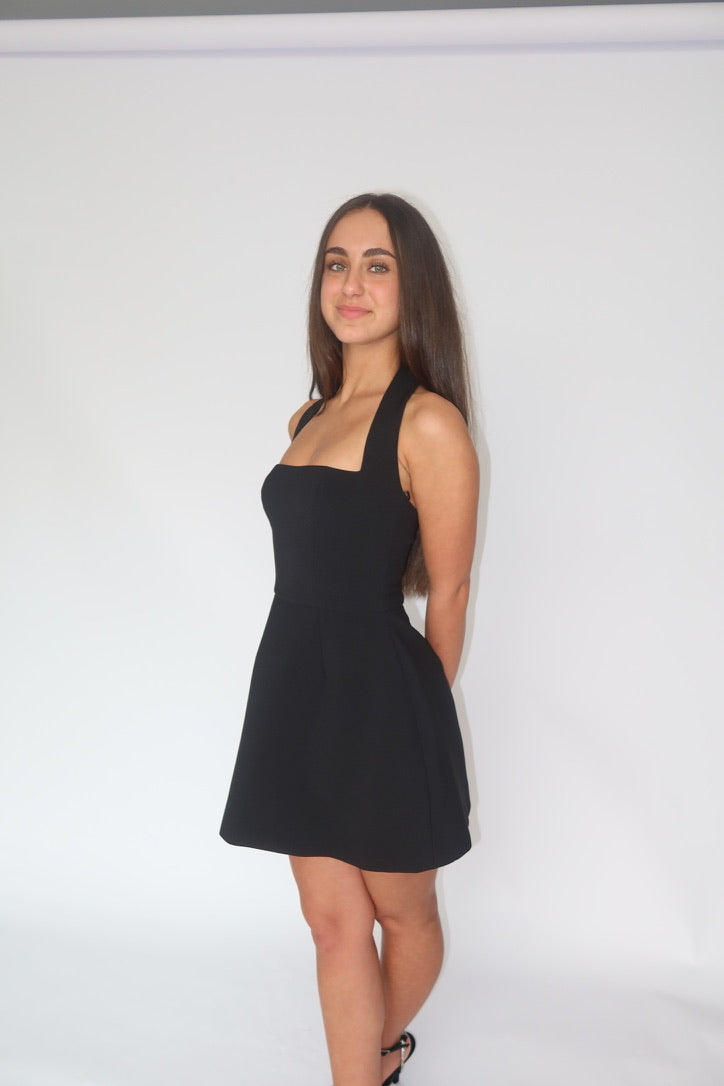 A woman modeling a black sleeveless mini dress with a square neckline and flared skirt, standing against a plain white studio background.