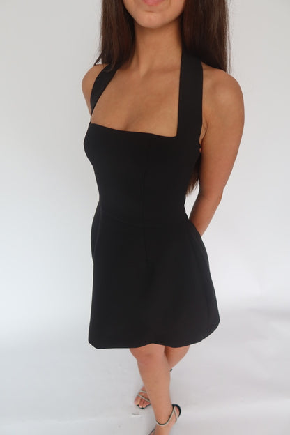 A woman wearing a black halter-neck mini dress with a square neckline, photographed against a plain white studio background.