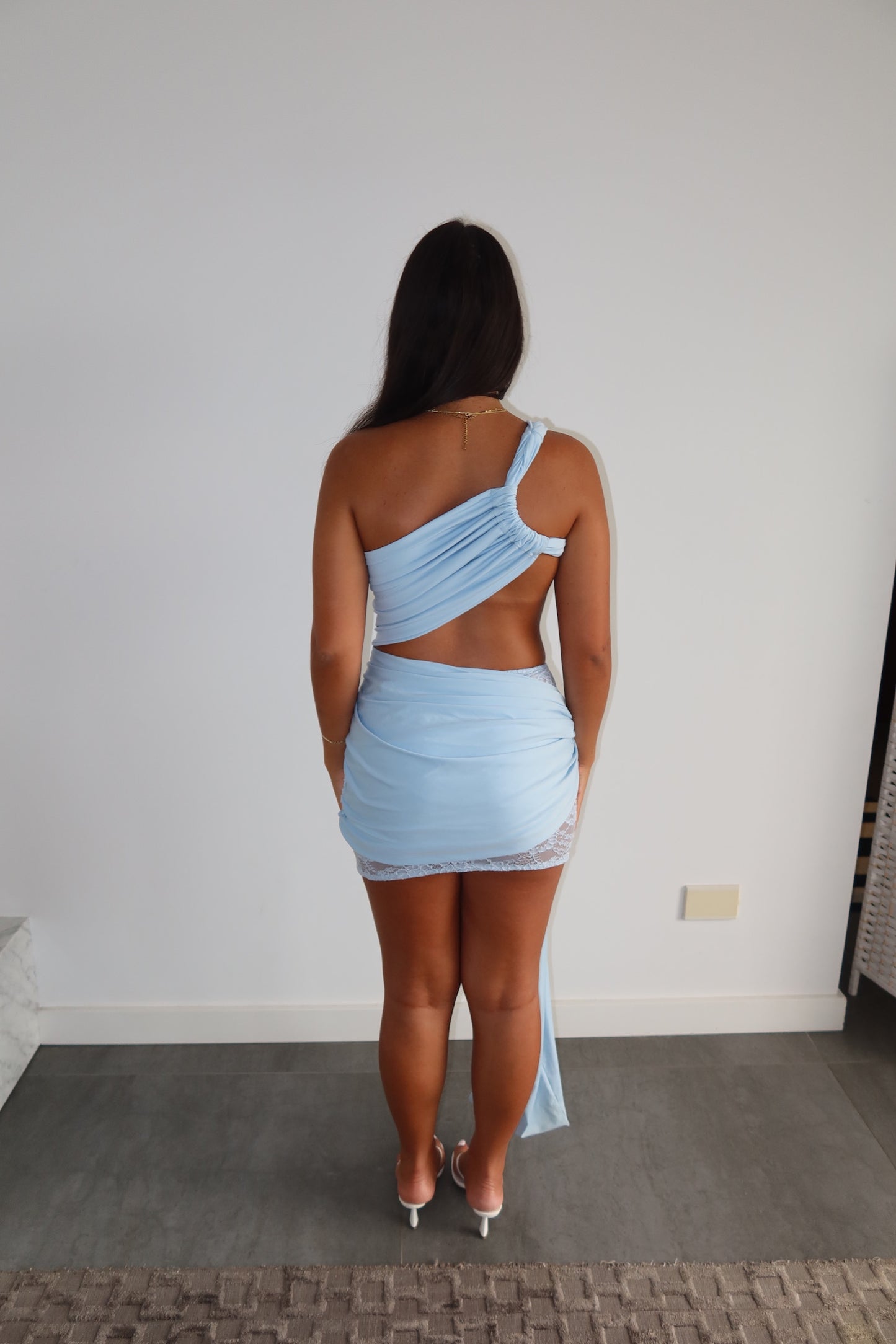 A woman viewed from behind wearing a light blue one-shoulder wrap mini dress.