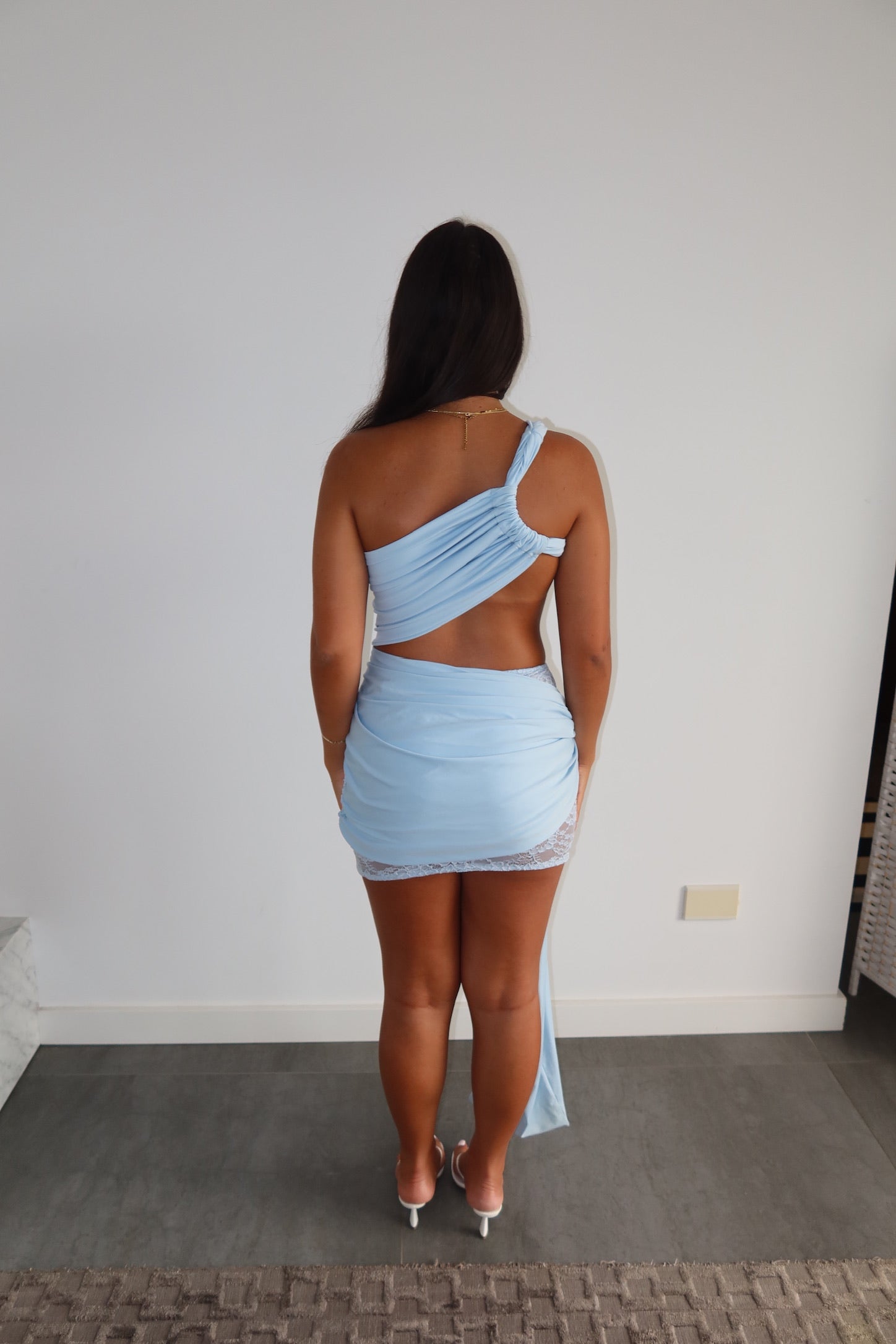 A woman viewed from behind wearing a light blue one-shoulder wrap mini dress.