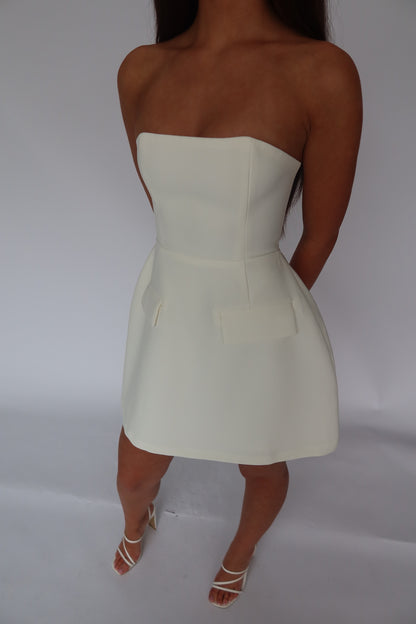 A woman modeling a strapless off-white mini dress with structured shoulders, a defined waist, and a flared skirt, shown against a plain light gray studio background.