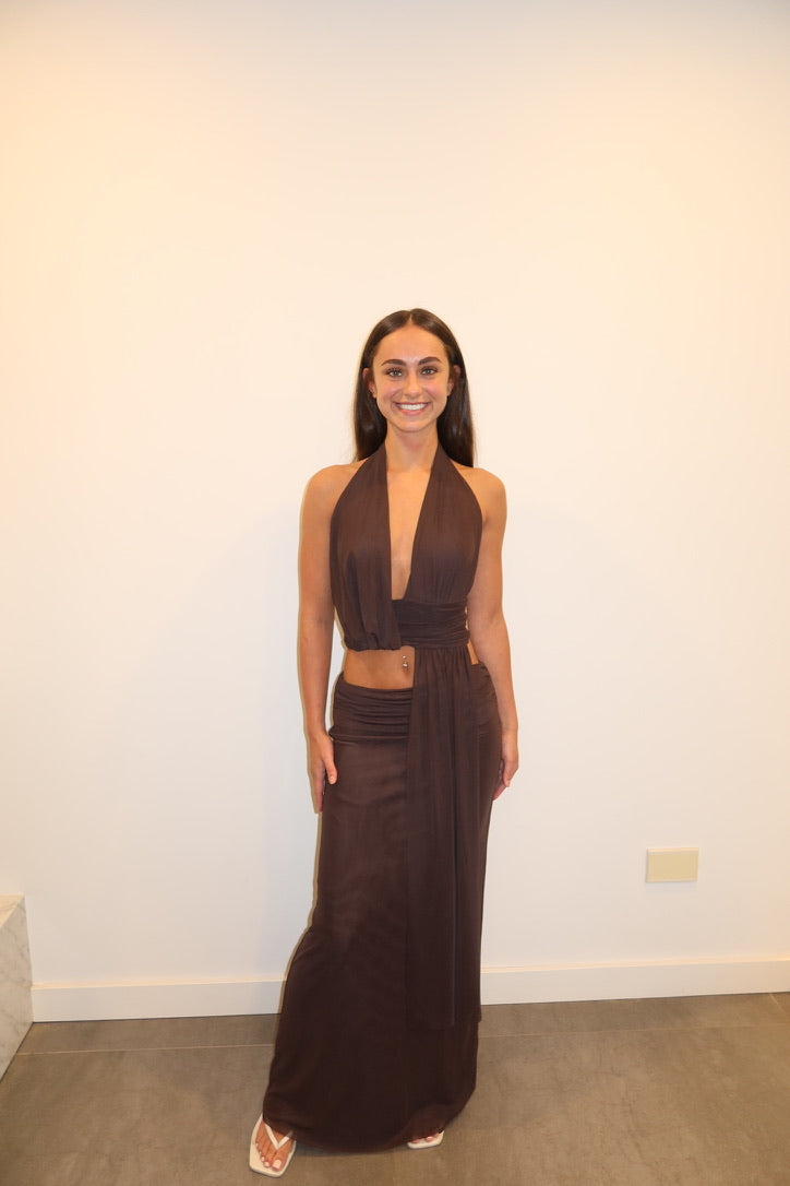 A woman modeling a dark brown two-piece halter dress set with a deep V-neck crop top and a matching draped maxi skirt, standing against a plain off-white wall.