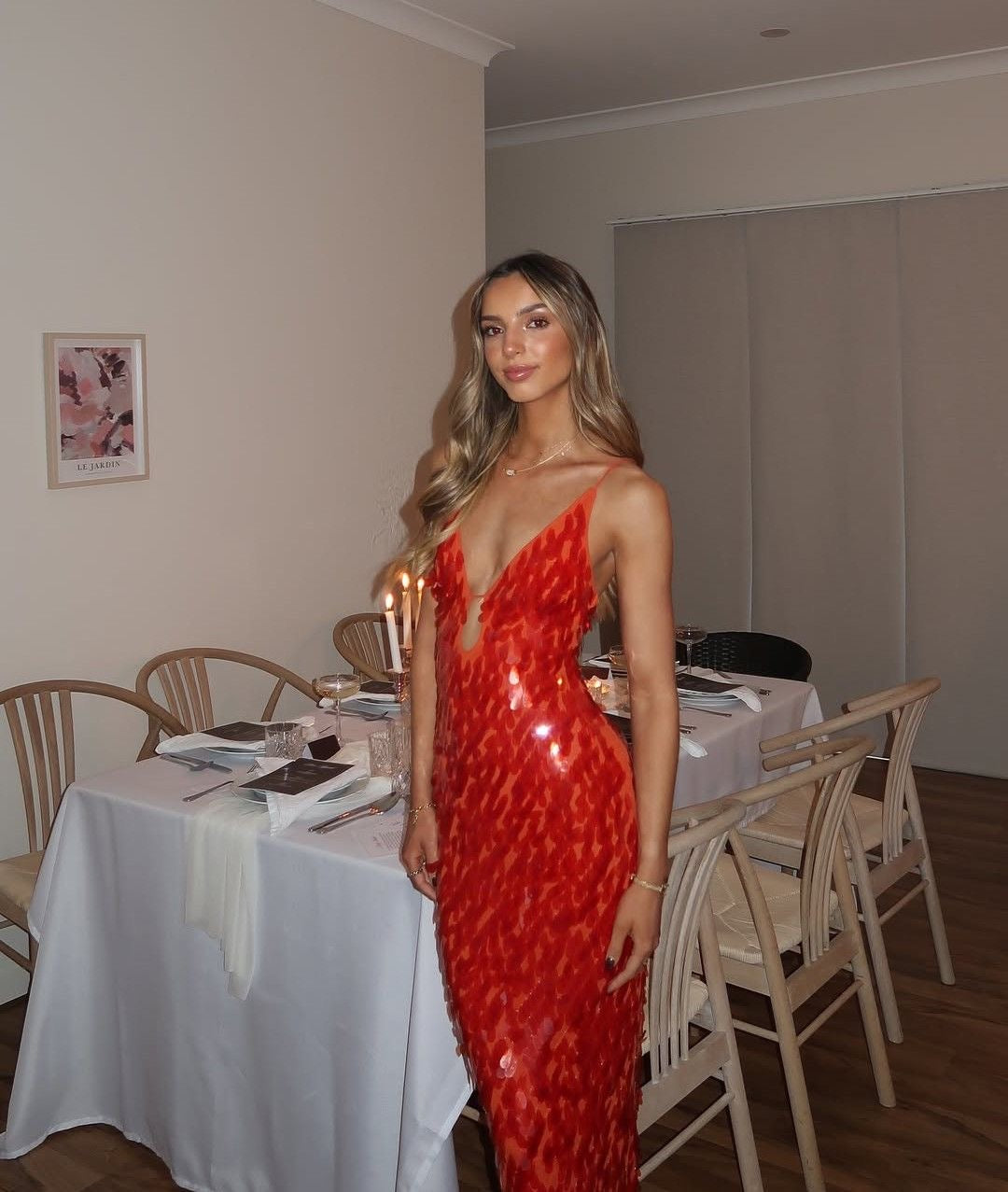 A woman wearing a red patterned spaghetti-strap evening gown stands in a dining room setting with a table and chairs behind her.
