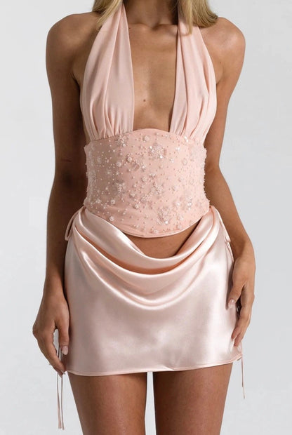 A model wearing a pale blush pink halter mini dress with a structured, beaded corset-style waist and draped satin skirt.