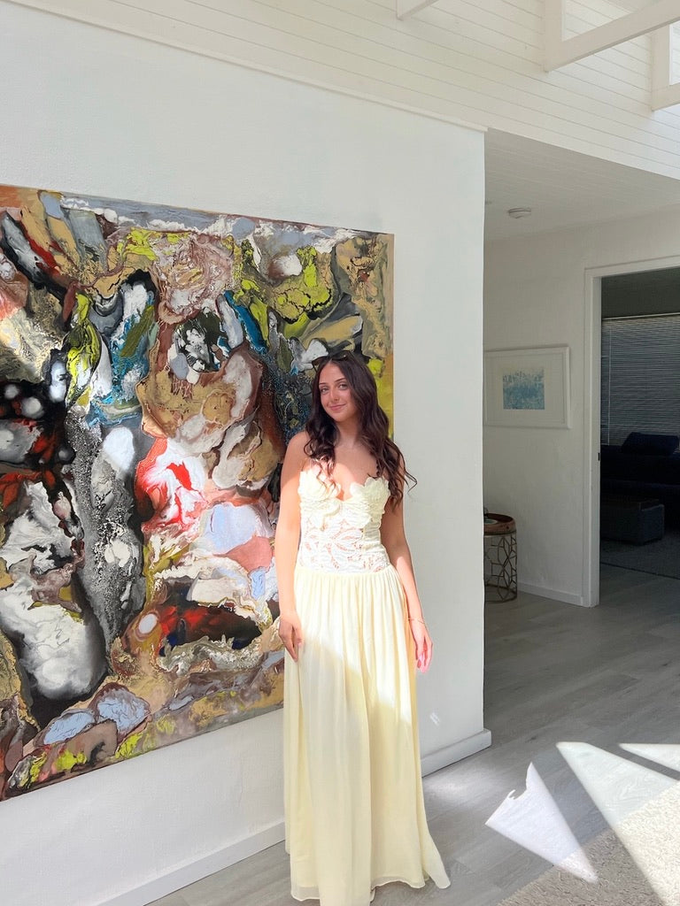 A woman wearing a cream-colored strapless maxi dress with a lace bodice stands in a bright room next to a large abstract painting.