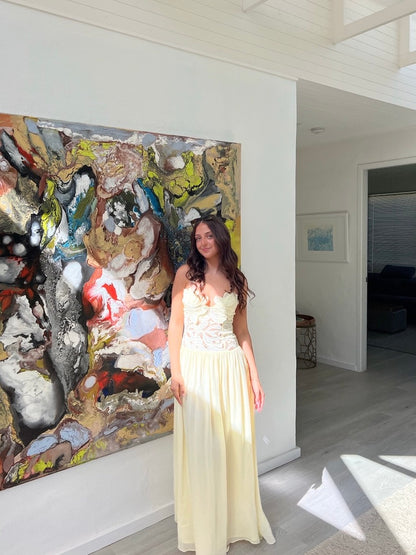 A woman wearing a cream-colored strapless maxi dress with a lace bodice stands in a bright room next to a large abstract painting.