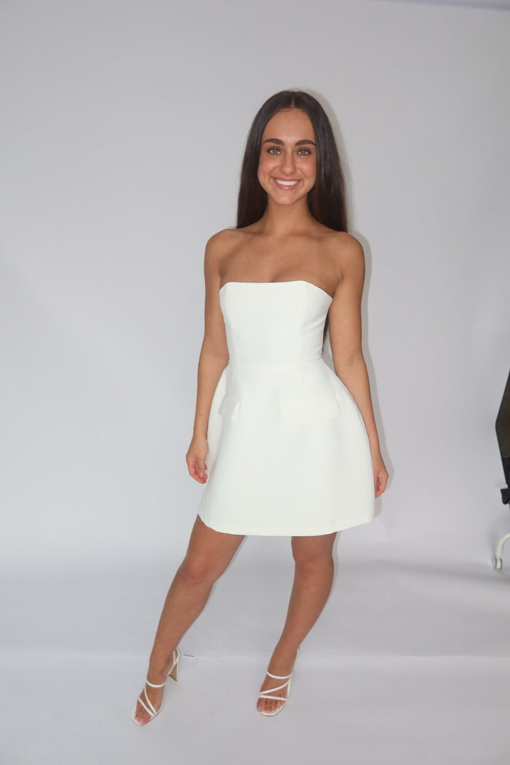 A woman modeling a strapless white mini dress with a flared skirt, paired with white strappy high-heeled sandals, set against a plain white studio background.
