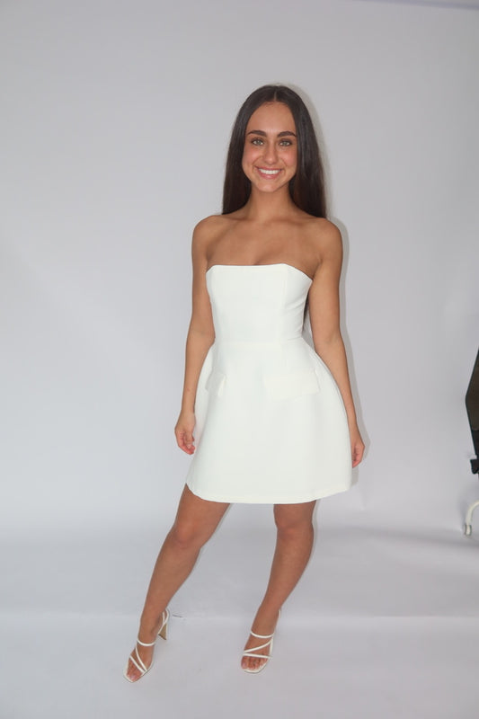 A woman modeling a strapless white mini dress with a flared skirt, paired with white strappy high-heeled sandals, set against a plain white studio background.