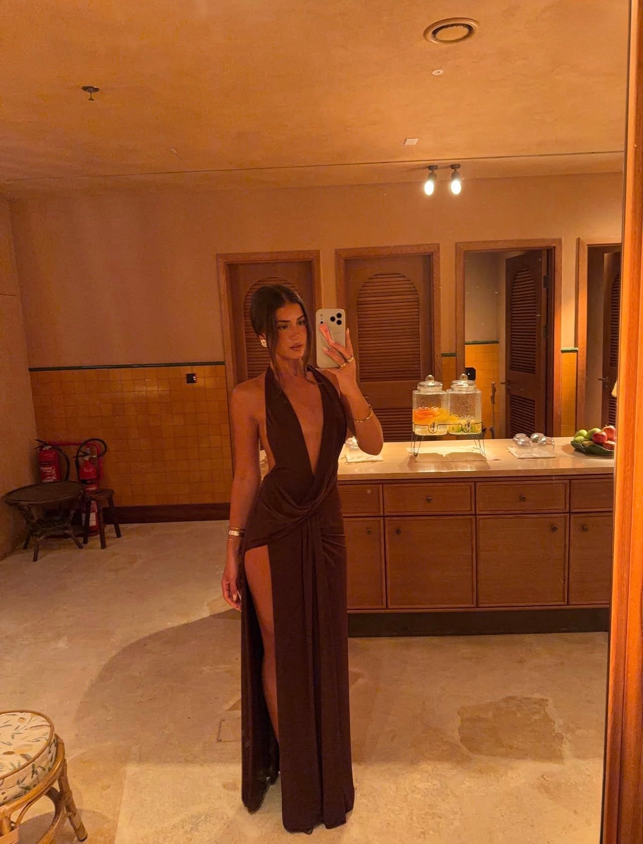 A woman takes a mirror selfie in a luxurious hotel-style bathroom wearing a dark brown halter-neck maxi dress with a deep plunging neckline and a high thigh slit.