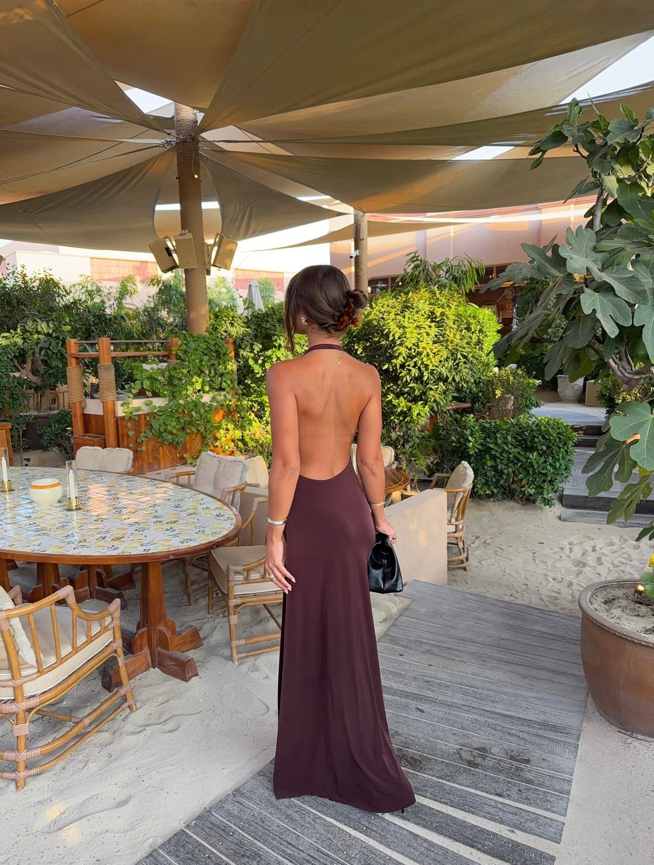 A woman viewed from behind wearing a floor-length open-back evening gown in a deep plum or burgundy-brown shade, standing on a patio under a large beige canopy with outdoor dining furniture and greenery around her.