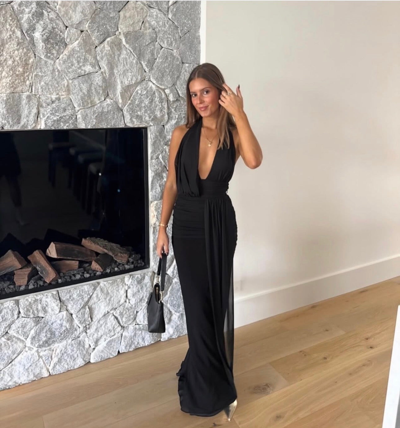 A woman wearing a black halter-neck maxi dress with a deep plunging neckline and a draped waist, standing in front of a stone fireplace and holding a small black handbag.