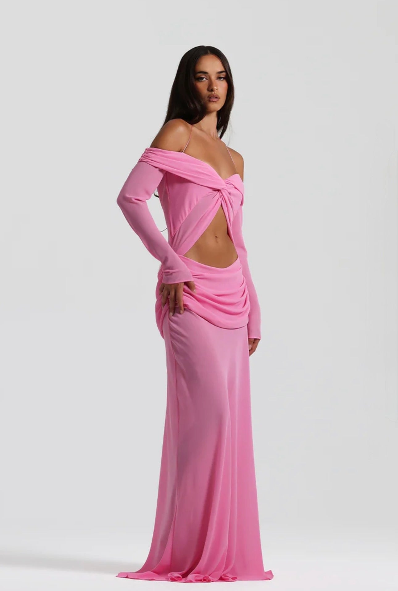 A woman modeling a floor-length off-the-shoulder pink gown with a twisted front detail, long sleeves, and a draped waist, photographed against a light gray studio background.
