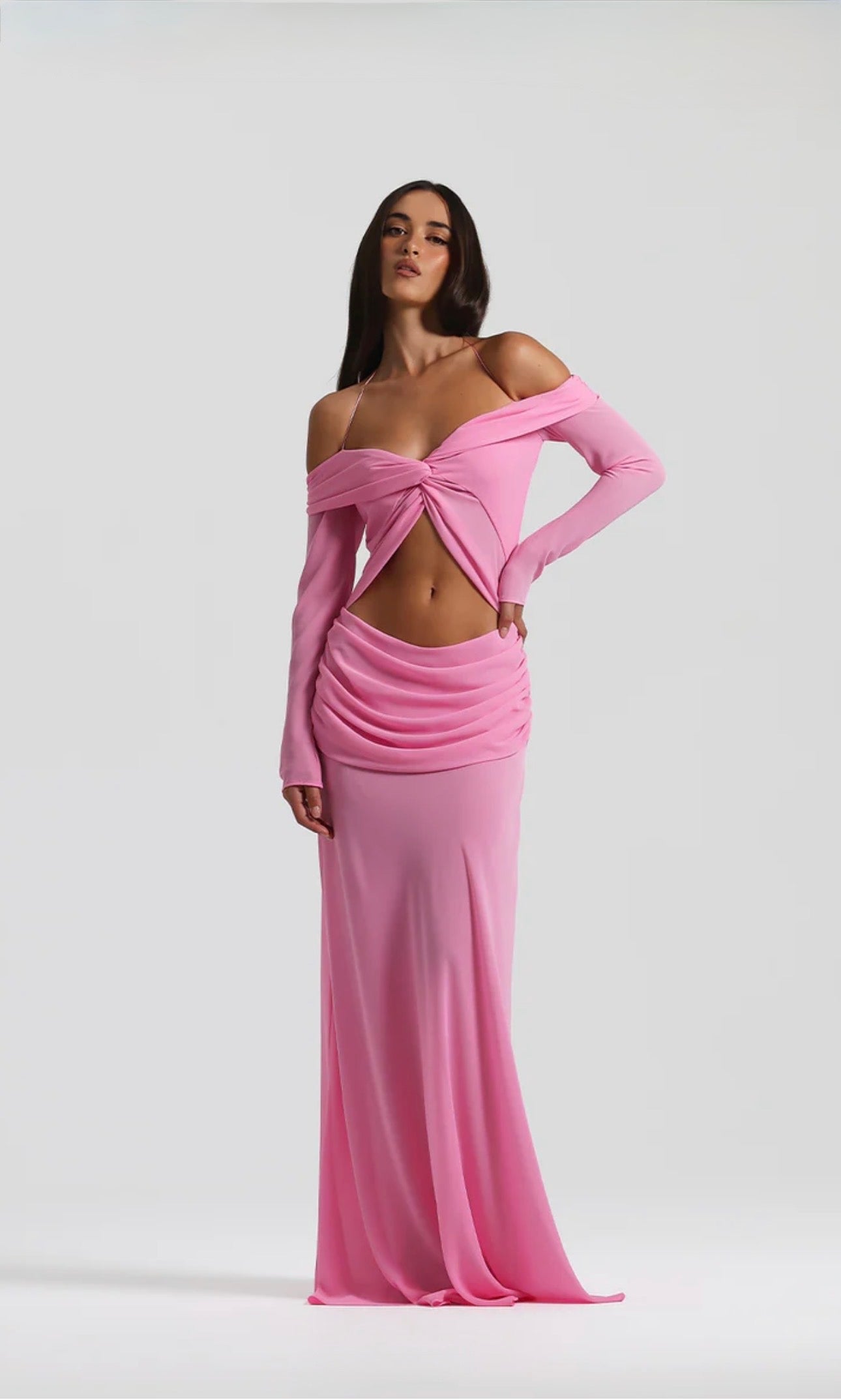 A model wearing a pink off-the-shoulder long-sleeve maxi dress with a twisted front detail and a ruched draped skirt, set against a plain light gray studio background.