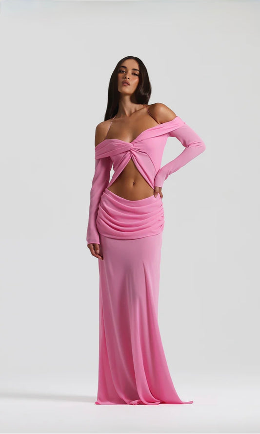 A model wearing a pink off-the-shoulder long-sleeve maxi dress with a twisted front detail and a ruched draped skirt, set against a plain light gray studio background.