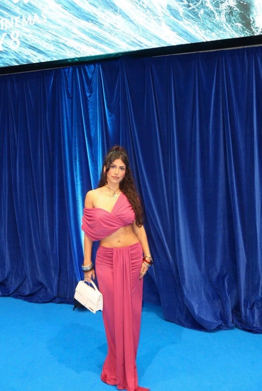 A woman stands on a blue carpet in front of a blue velvet curtain backdrop, wearing a bright pink two-piece outfit with an asymmetrical draped top and flowing skirt. She is holding a small white handbag.