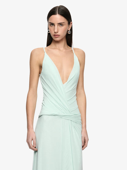 A woman modeling a mint green sleeveless wrap dress with a deep V-neckline and draped waist details, set against a plain light gray background.