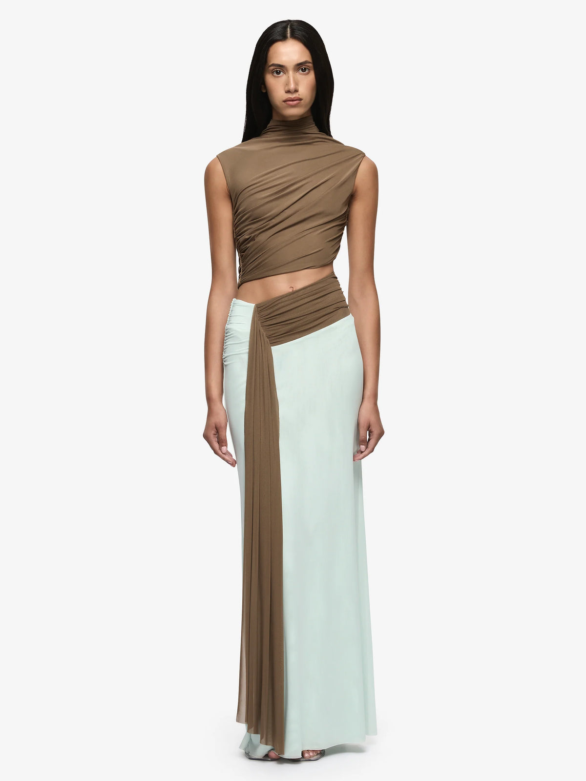 A model wearing a two-piece draped outfit consisting of a brown mock-neck cropped top and a matching pleated skirt in brown and pale mint green, shot against a solid light gray background.