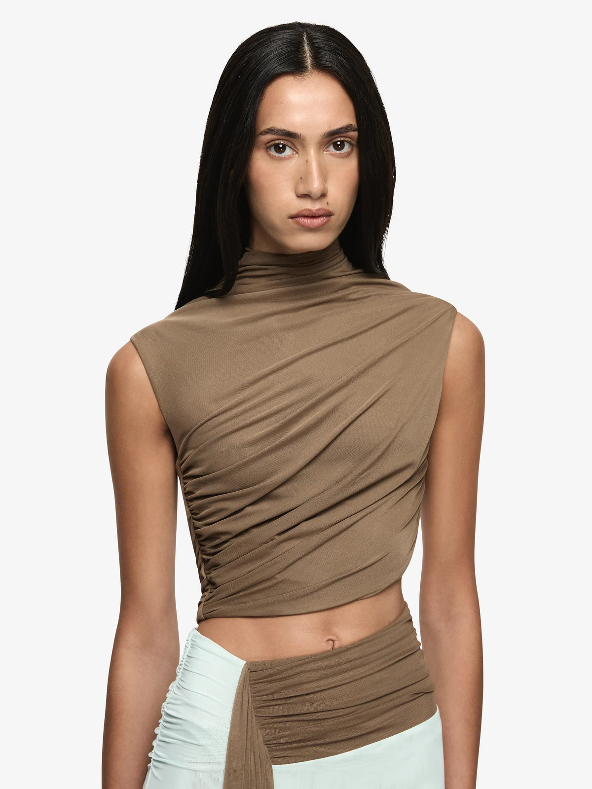A model wearing a sleeveless, high-neck, ruched crop top in taupe brown, paired with a light mint green wrap skirt, against a plain light gray background.