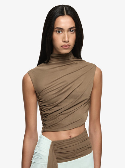 A model wearing a sleeveless, high-neck, ruched crop top in taupe brown, paired with a light mint green wrap skirt, against a plain light gray background.