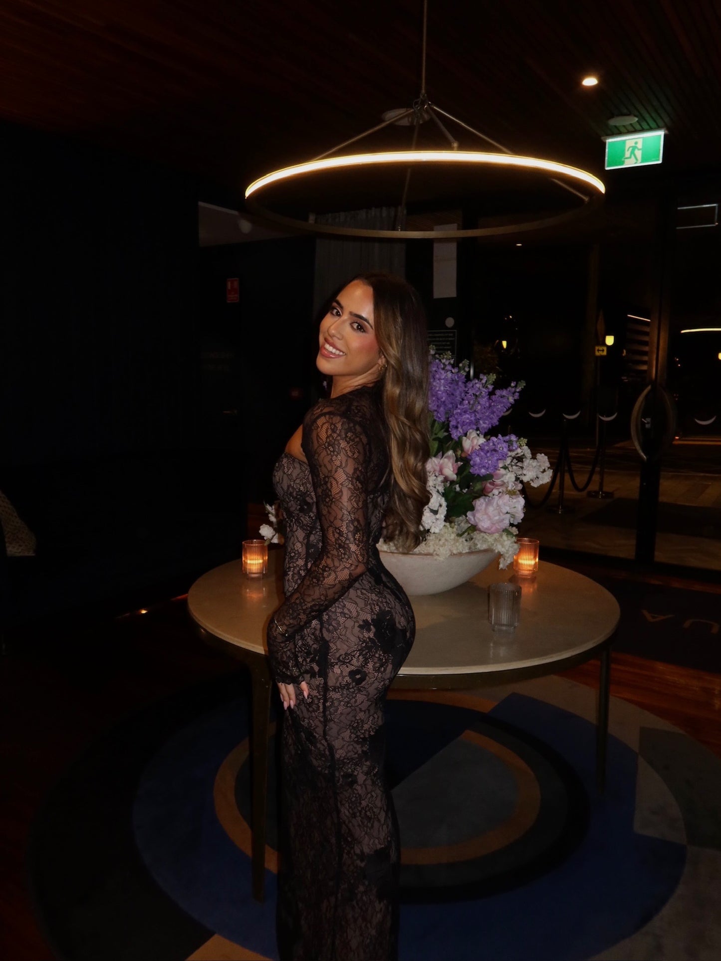 A woman wearing a black lace long-sleeve maxi dress stands beside a round table with a floral arrangement in a dimly lit indoor venue.