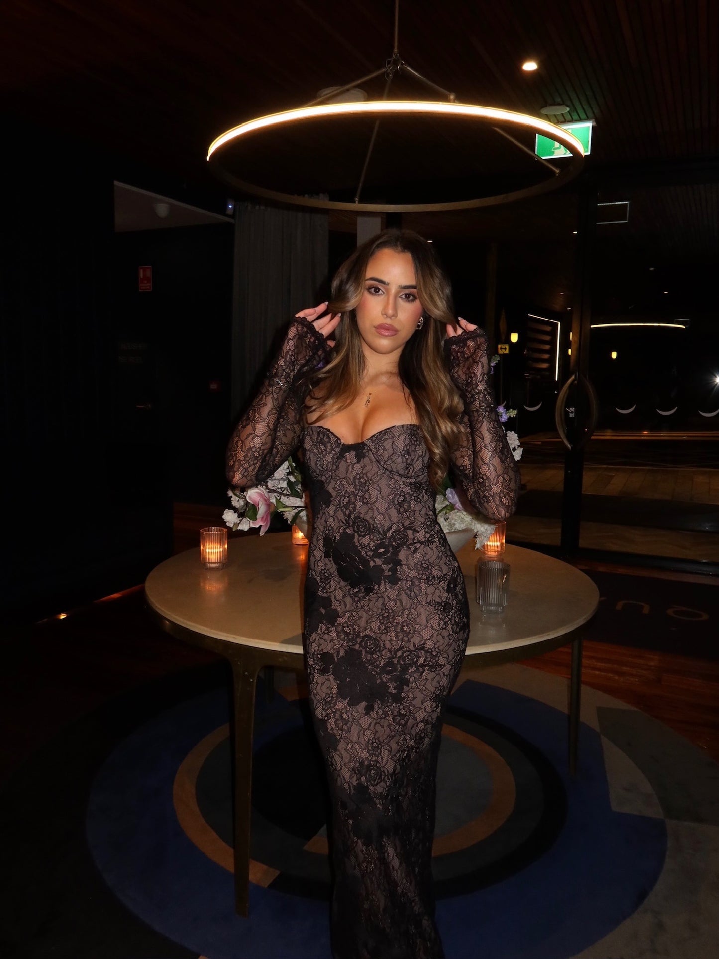 A woman wearing a sheer black lace long-sleeve gown with a sweetheart neckline, standing in a dimly lit indoor venue with a round table and candles behind her.