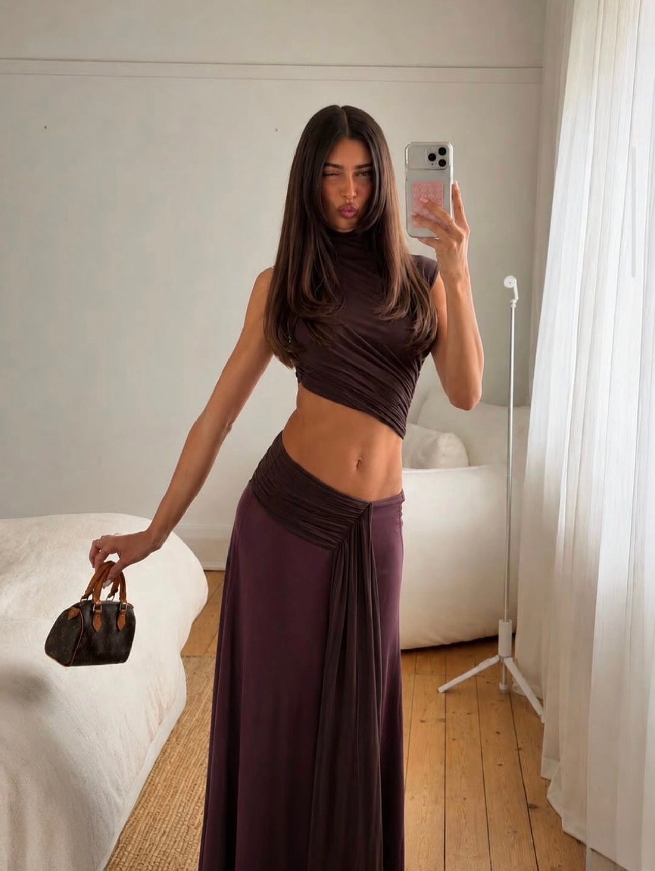 A woman takes a mirror selfie in a bedroom wearing a dark plum purple two-piece set consisting of a ruched crop top and a matching long draped skirt. She is holding a small dark brown handbag.