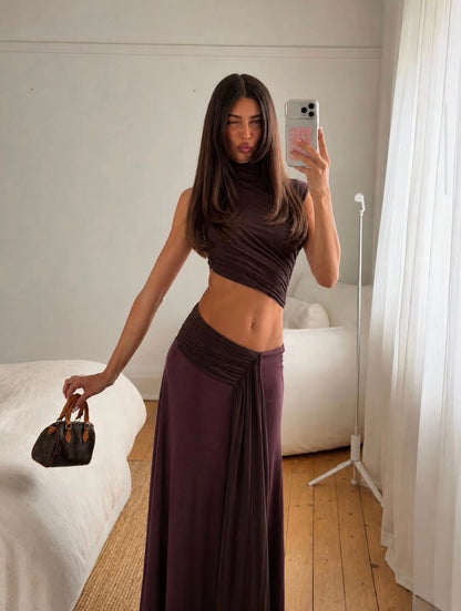 A woman takes a mirror selfie in a bedroom wearing a dark plum purple two-piece set consisting of a ruched crop top and a matching long draped skirt. She is holding a small dark brown handbag.