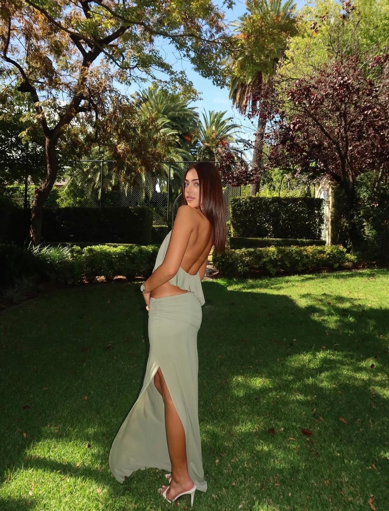 A woman stands on a sunny grassy lawn in a garden, wearing a light sage green backless maxi dress with a thigh-high slit and white high-heeled sandals.