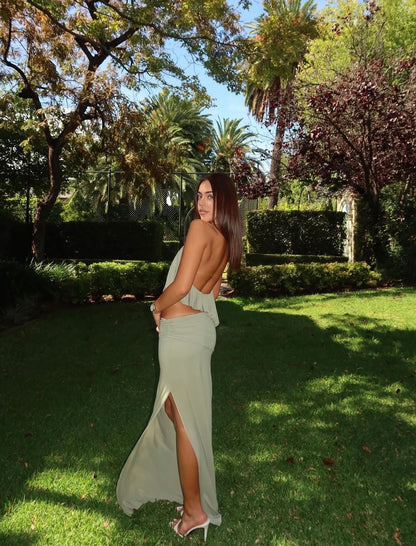 A woman stands on a sunny grassy lawn in a garden, wearing a light sage green backless maxi dress with a thigh-high slit and white high-heeled sandals.