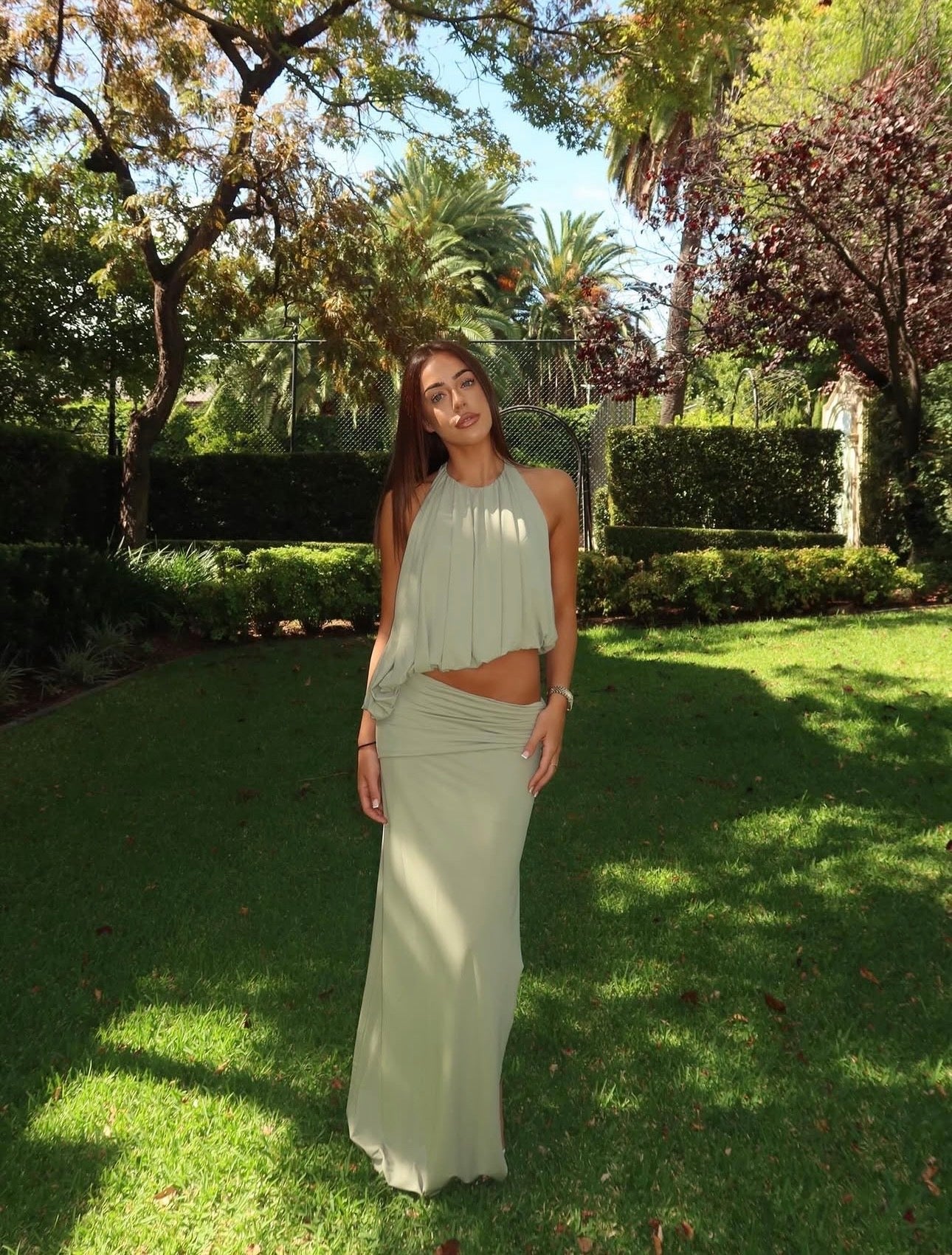 A woman wearing a light sage green two-piece outfit consisting of a sleeveless pleated crop top and a matching high-waisted maxi skirt, standing in a sunny garden with green grass and trees.