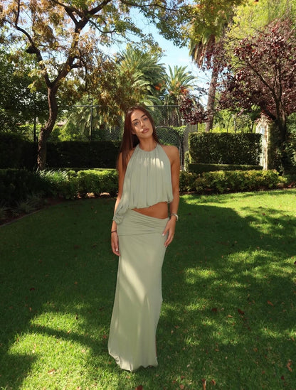 A woman wearing a light sage green two-piece outfit consisting of a sleeveless pleated crop top and a matching high-waisted maxi skirt, standing in a sunny garden with green grass and trees.