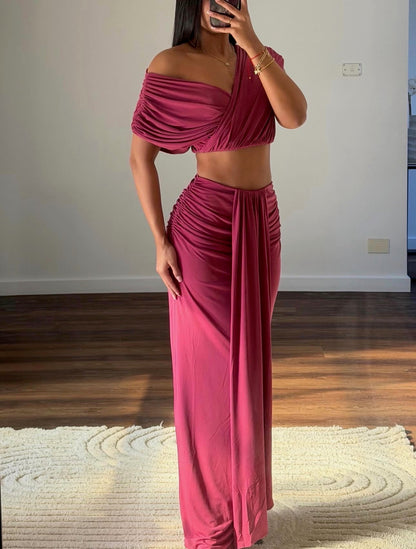 A woman takes a mirror selfie wearing a matching two-piece outfit in a dusty rose or magenta color. The set includes an off-the-shoulder draped crop top and a high-waisted, gathered maxi skirt.