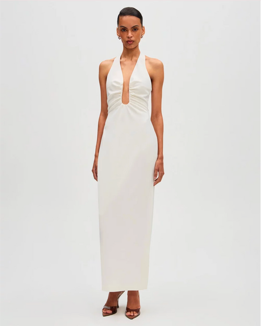 A woman modeling a sleeveless white halter-neck maxi dress with a deep plunging neckline and gathered waist details, set against a plain light gray studio background.