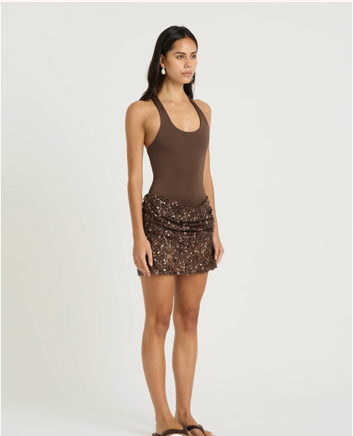 A woman modeling a sleeveless brown bodysuit with a ruched, sequined mini skirt against a plain light gray studio background.