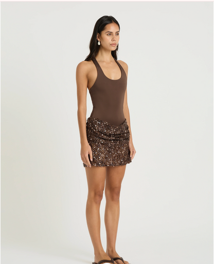 A woman modeling a sleeveless brown bodysuit with a ruched, sequined mini skirt against a plain light gray studio background.