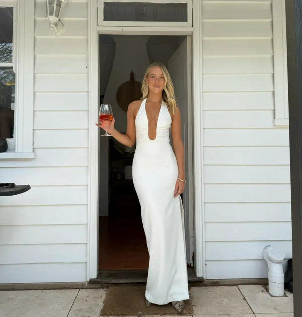 A woman stands in a doorway wearing a fitted white sleeveless gown with a deep plunging neckline and a floor-length silhouette. She holds a wine glass in one hand, with a white house exterior behind her.