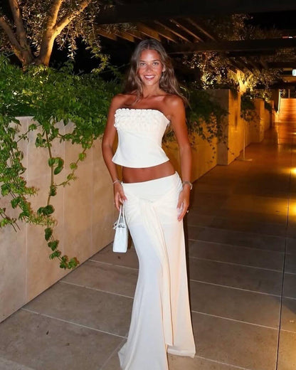 A woman stands outdoors at night wearing a white two-piece outfit consisting of a strapless cropped top with textured detailing and a matching draped maxi skirt. She is holding a small white handbag, with warm lights illuminating a walkway and greenery on the left.