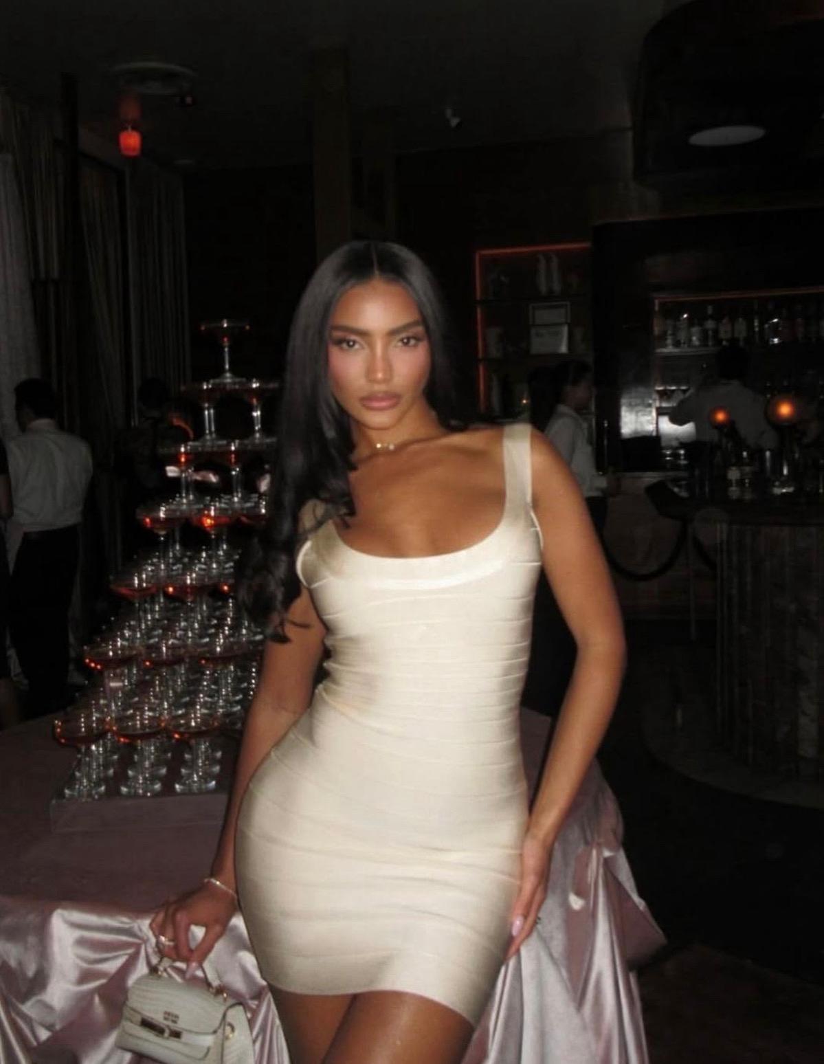 A woman posing in a dimly lit indoor venue wearing a fitted, ruched off-white mini dress with a scoop neckline.