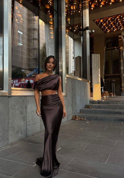 A woman models a dark brown satin two-piece set consisting of an off-the-shoulder crop top and a matching high-waisted flared skirt, standing on a stone walkway outside a modern building.