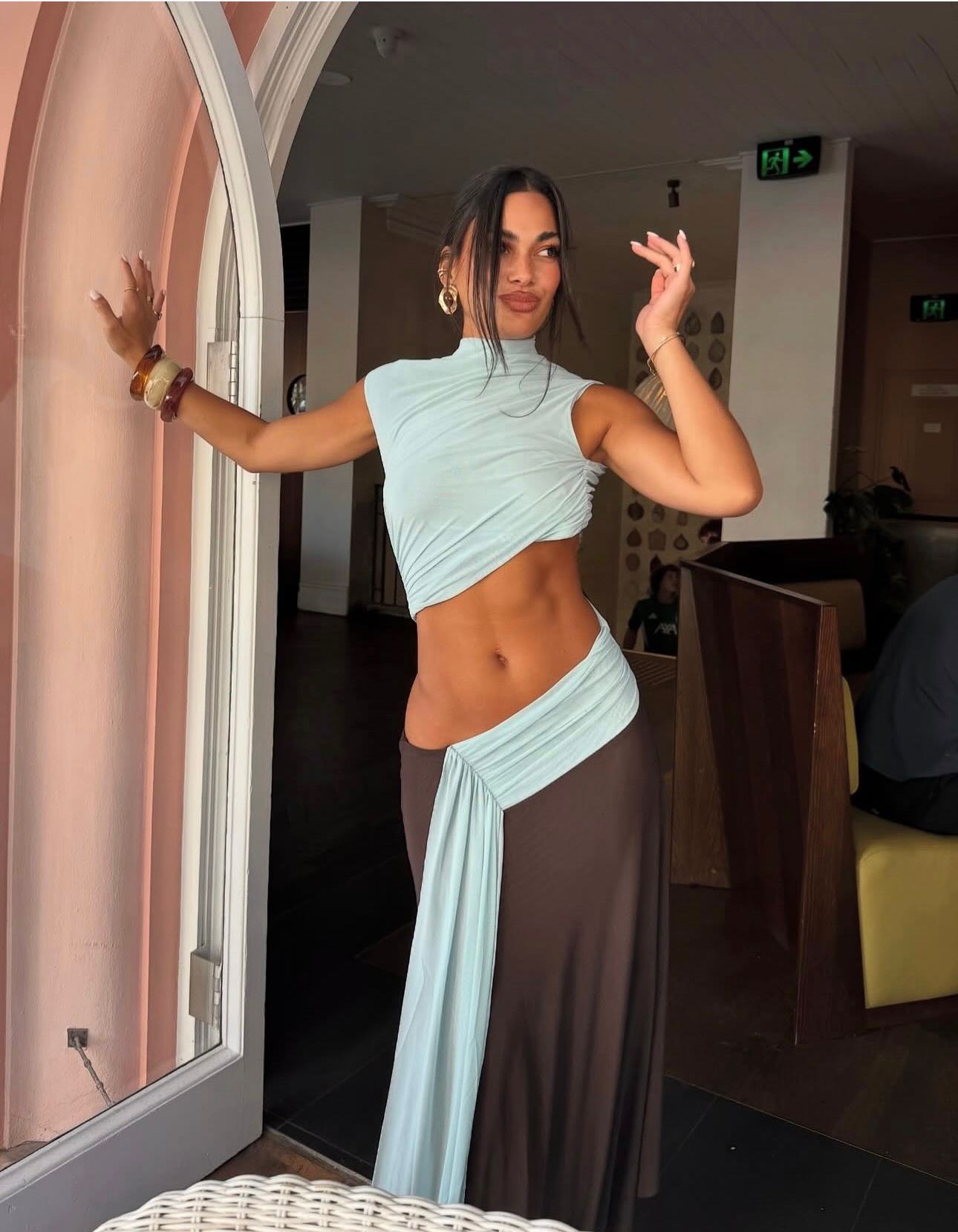 A woman posing indoors in a light blue wrap-style crop top and matching draped skirt, with a dark brown long skirt layer underneath, standing near a pink arched doorway.